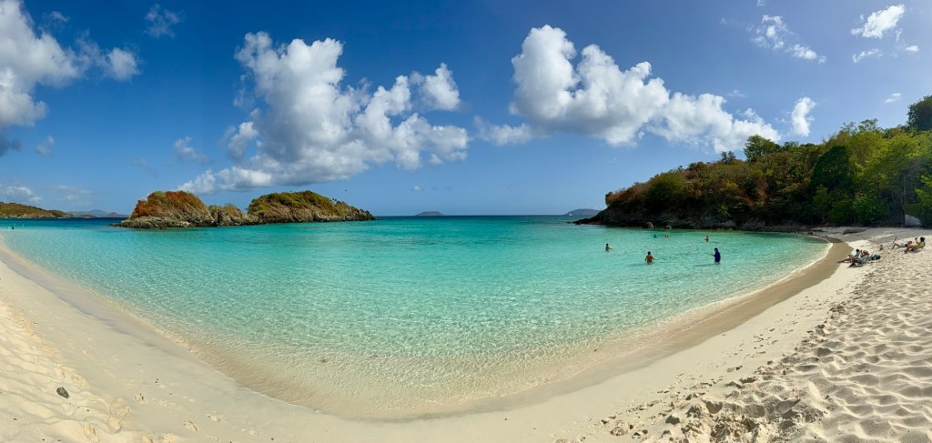 A panoramic view of a serene beach with white sand, clear turquoise waters, and scattered rocky islands under a blue sky dotted with fluffy white clouds. Some people are enjoying the water while others relax on the shore.