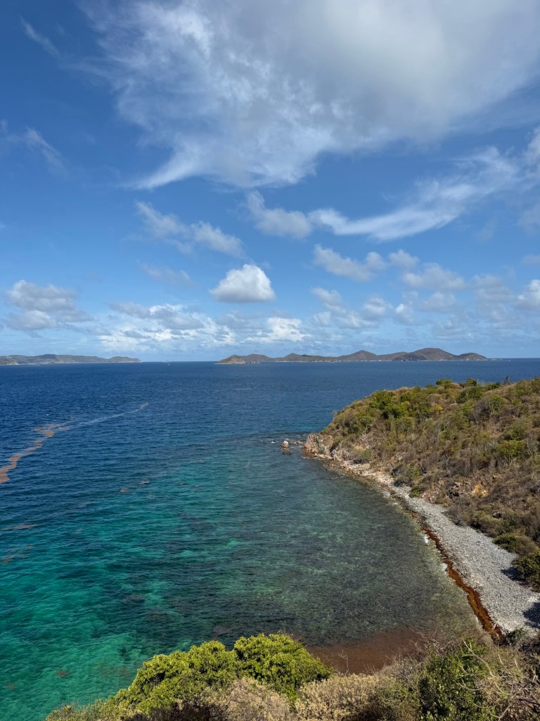 A panoramic view of a coastal landscape featuring clear turquoise waters, rocky shores, and lush greenery under a partly cloudy sky.