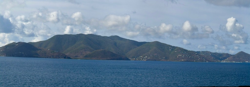 A panoramic view of green hills rising from the blue ocean under a cloudy sky.