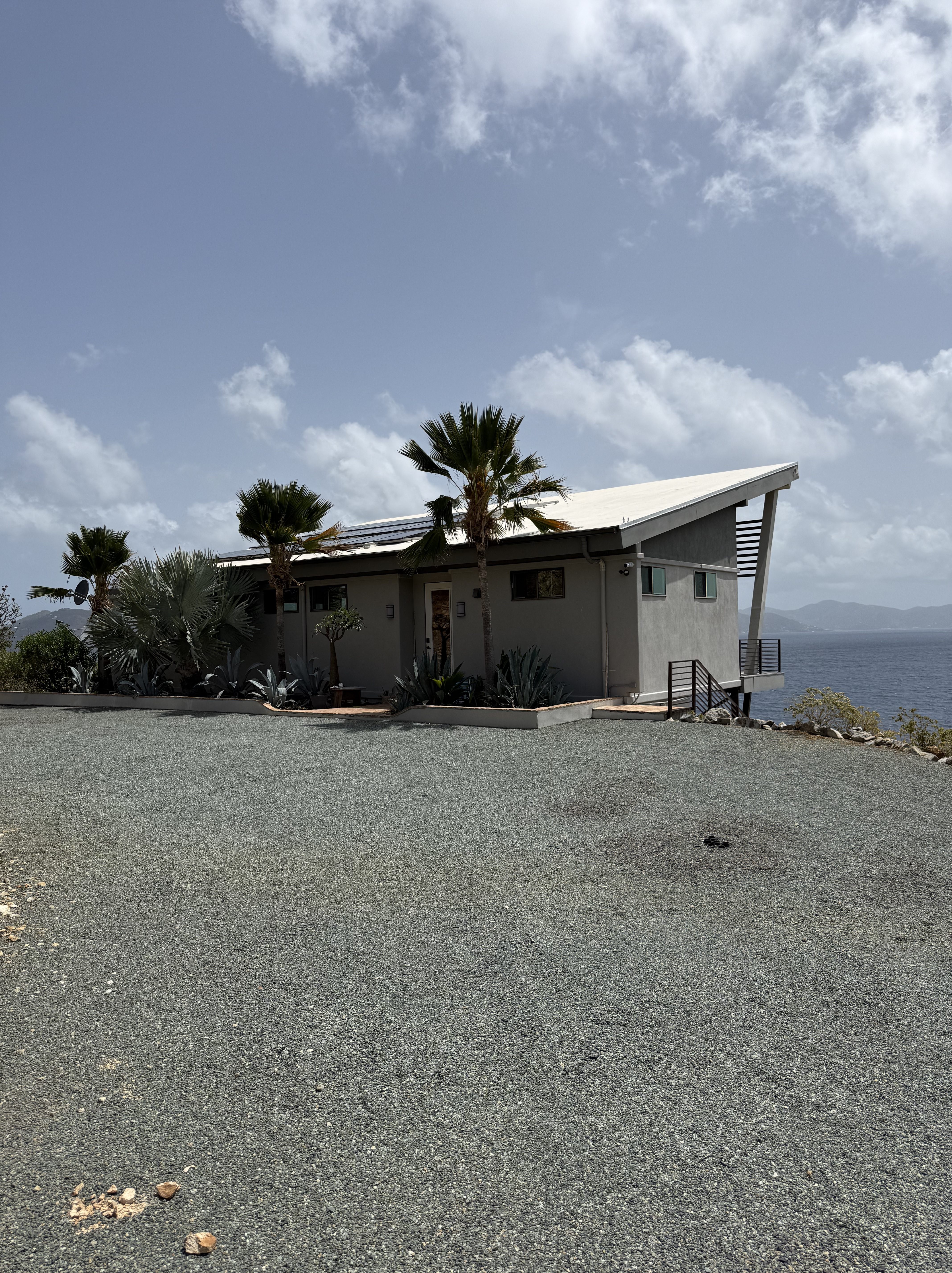 A modern house with a sloped roof surrounded by palm trees and desert plants, located near the water under a cloudy sky.