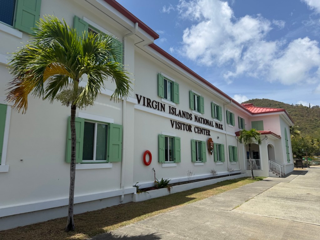 Exterior view of the Virgin Islands National Park Visitor Center featuring a white building with green shutters and a red roof, surrounded by palm trees and blue skies.