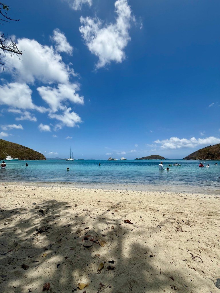 A scenic beach view with clear blue water, gentle waves, and a sandy shore, featuring people swimming and enjoying the sun under a bright sky with scattered clouds.