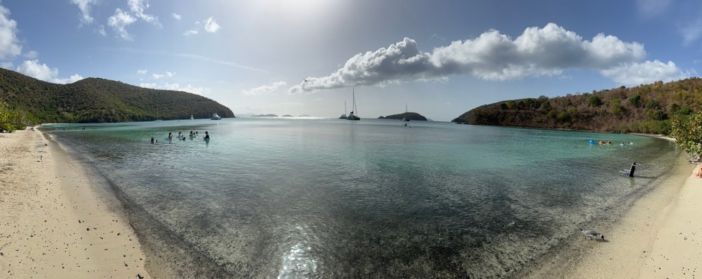 A panoramic view of a serene beach with clear turquoise water, featuring people swimming and enjoying the sun, surrounded by lush green hills and distant islands.