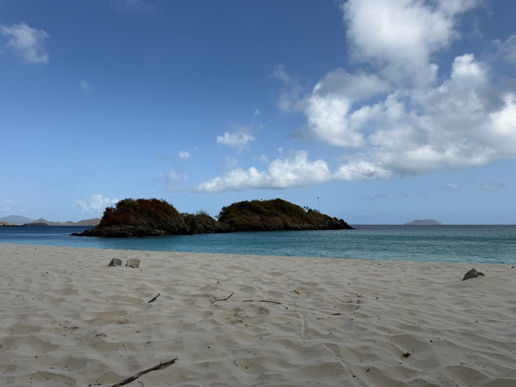 Scenic beach view with soft sand, turquoise water, and small rocky islands under a blue sky with clouds.